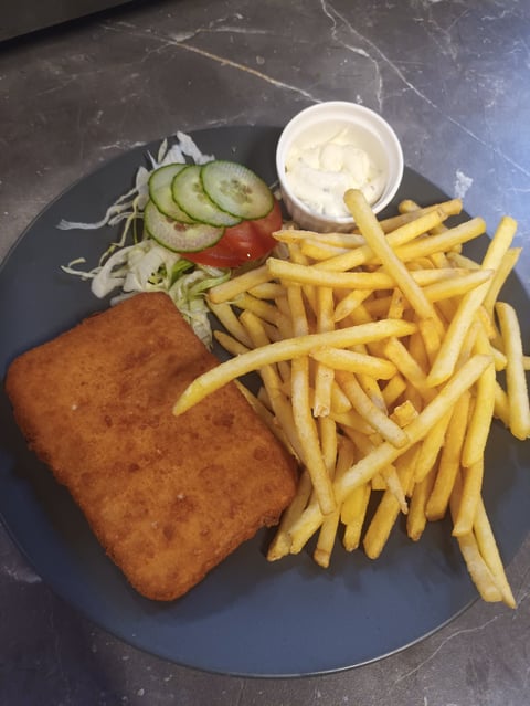 A plate with a fried fish fillet, golden french fries, coleslaw, tomato and cucumber slices, and a small bowl of white sauce